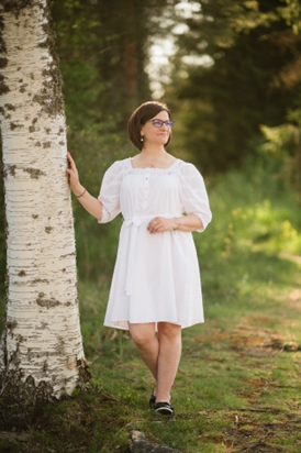 Valokuva Salla Hewittista. Salla nojaa kesäiltana koivunrunkoon, katselee luontoa ja hymyilee.